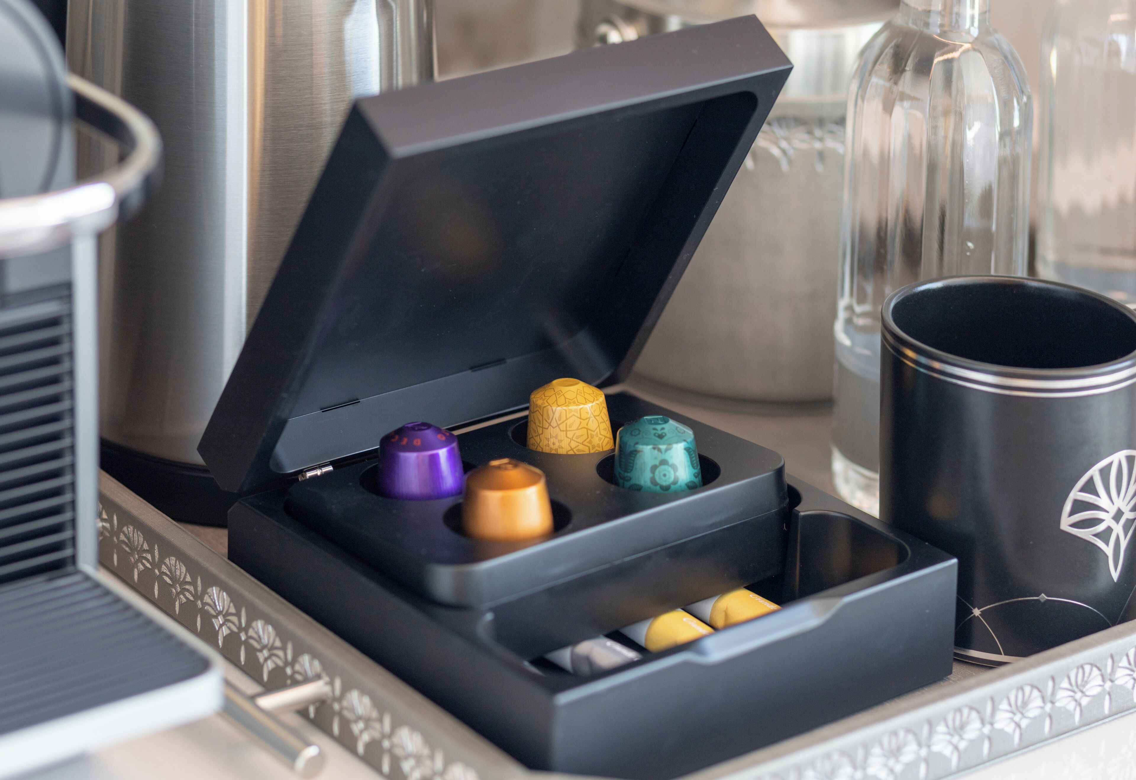 Un recipiente negro con varios compartimentos que contienen cápsulas de colores, una taza negra y una computadora portátil sobre una mesa.