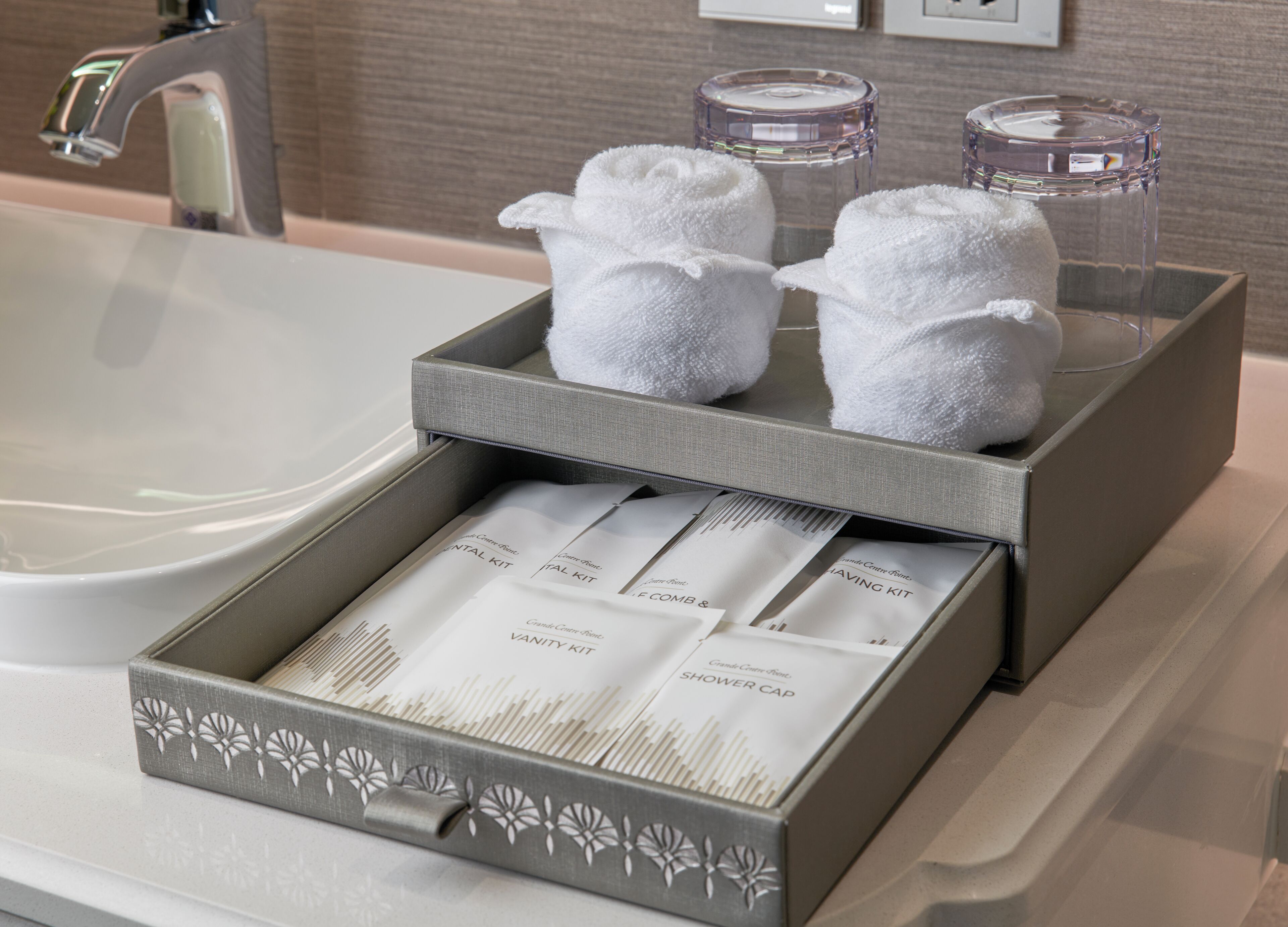 A bathroom with a grey towel rack holding two white towels, a glass container, and a box with toiletries.