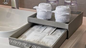 A bathroom with a grey towel rack holding two white towels, a glass container, and a box with toiletries.