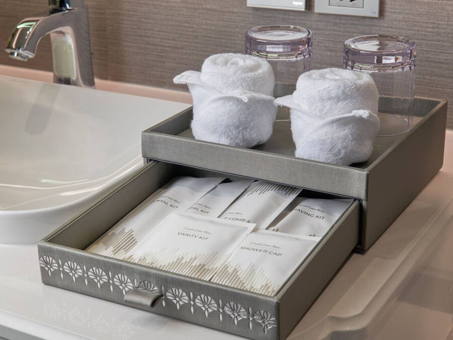 A bathroom with a grey towel rack holding two white towels, a glass container, and a box with toiletries.