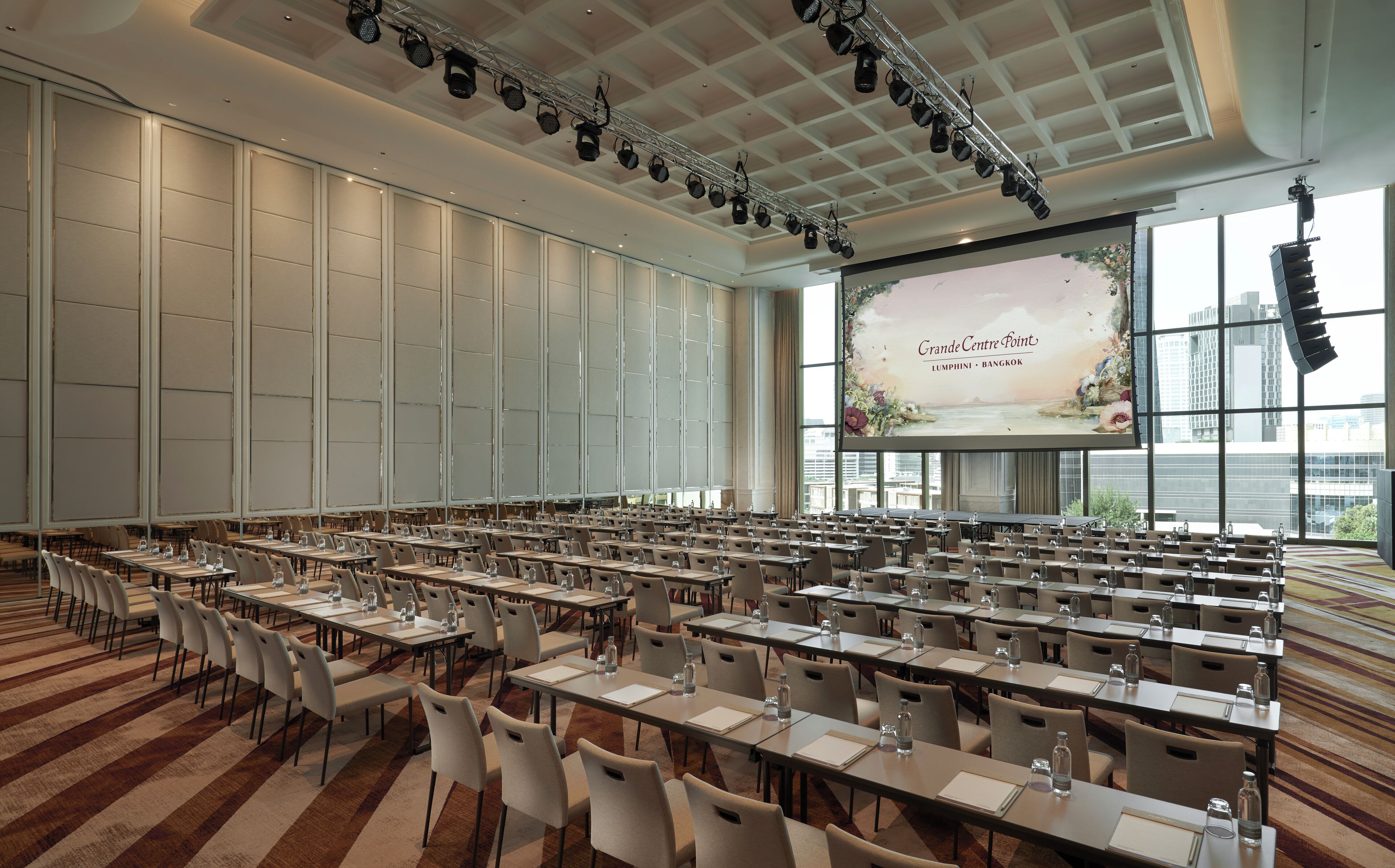 A conference room with rows of tables and chairs, large windows, and a stage with a presentation screen.