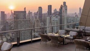 A rooftop bar with a city view at sunset, featuring wicker furniture and a glass railing.