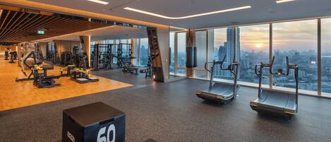 Un gimnasio moderno con diversos equipos de ejercicio, un amplio ventanal con vistas a la ciudad y una caja negra con el número 60 en el suelo.