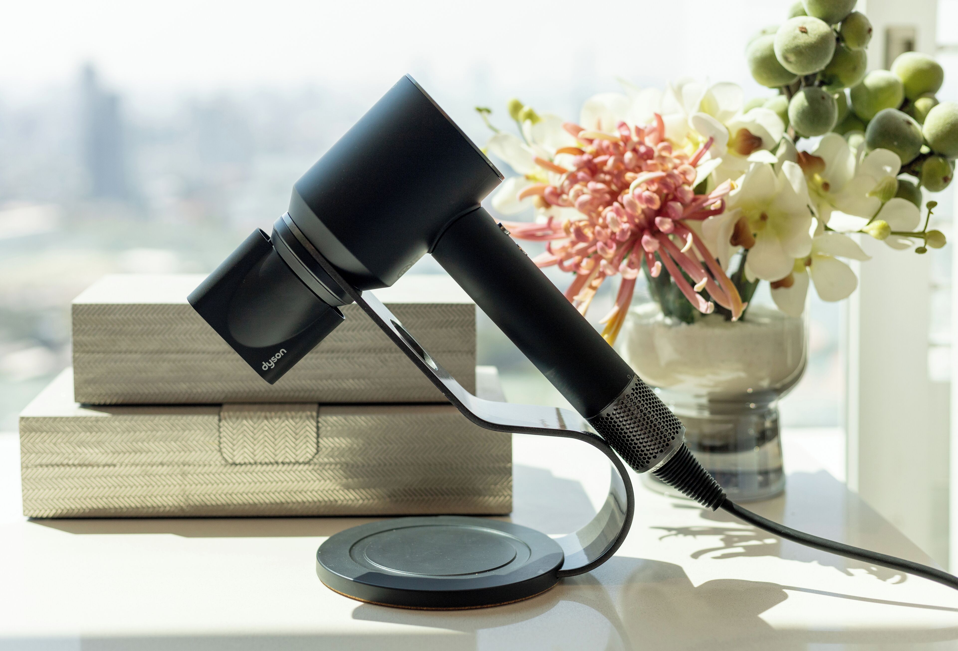 A Dyson hair dryer on a table with a vase of flowers and a book in the background.