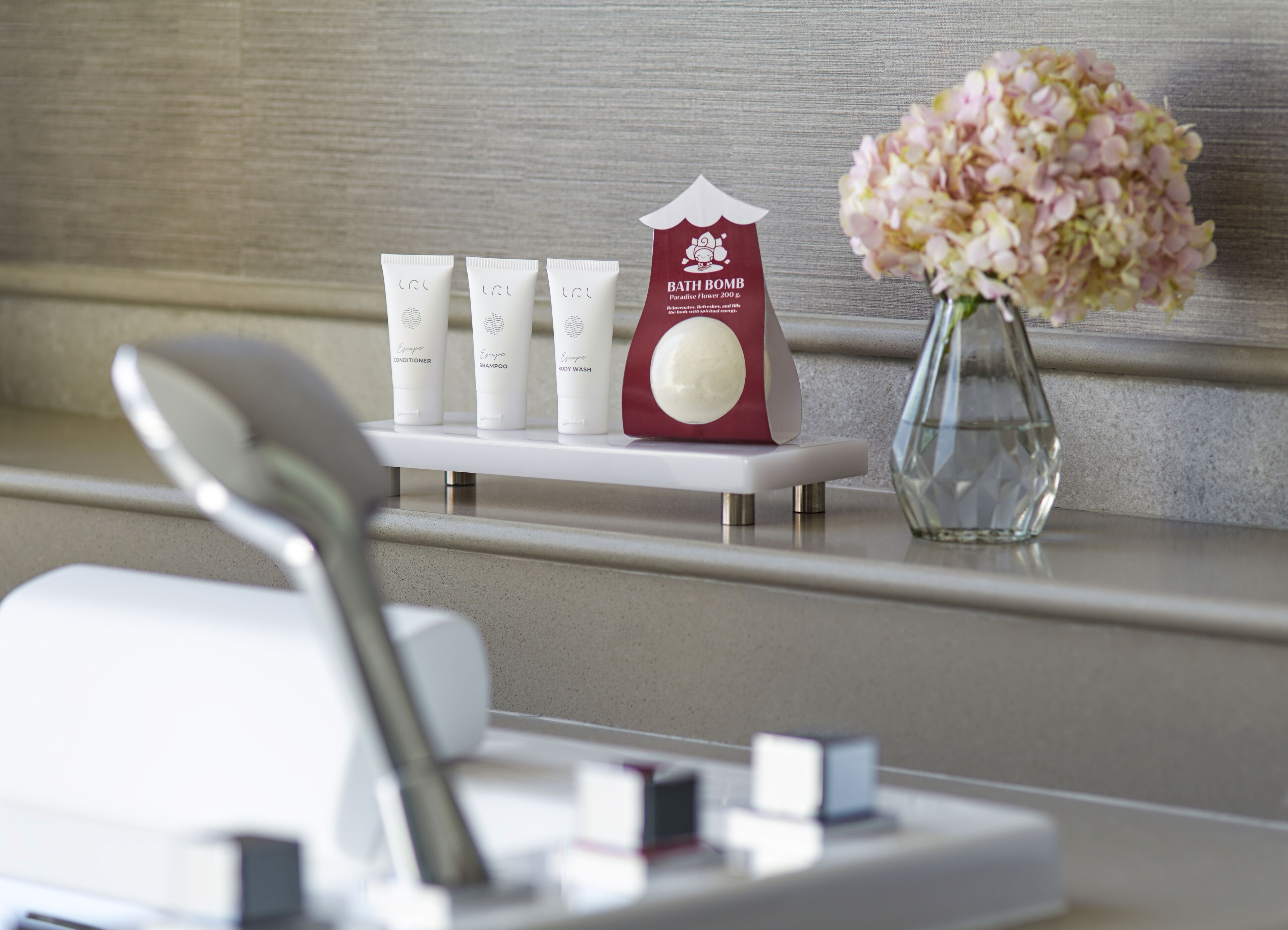 A bathroom vanity with a white sink, a mirror, and a shelf holding toiletries including a red bath bomb, three white tubes of shampoo, and a vase with pink flowers.
