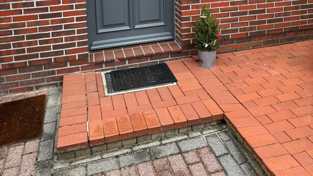 A brick building with a dark grey door, a small potted plant, and a metal doormat. - Apartment 'Ahlers' with Shared Terrace, Private Garden and Wi-Fi (Edewecht)