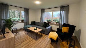 A modern living room with a sofa, a coffee table, a chair, and a desk. There are large windows with curtains, potted plants, and a view of trees and houses. - Apartment 'Ahlers' with Shared Terrace, Private Garden and Wi-Fi (Edewecht)