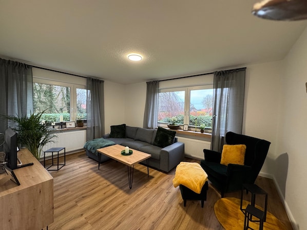 A modern living room with a sofa, a coffee table, a chair, and a desk. There are large windows with curtains, potted plants, and a view of trees and houses.