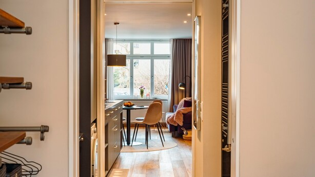Un pasillo que conduce a un comedor con una mesa y sillas, una ventana con cortinas, y una cama con edredón púrpura.