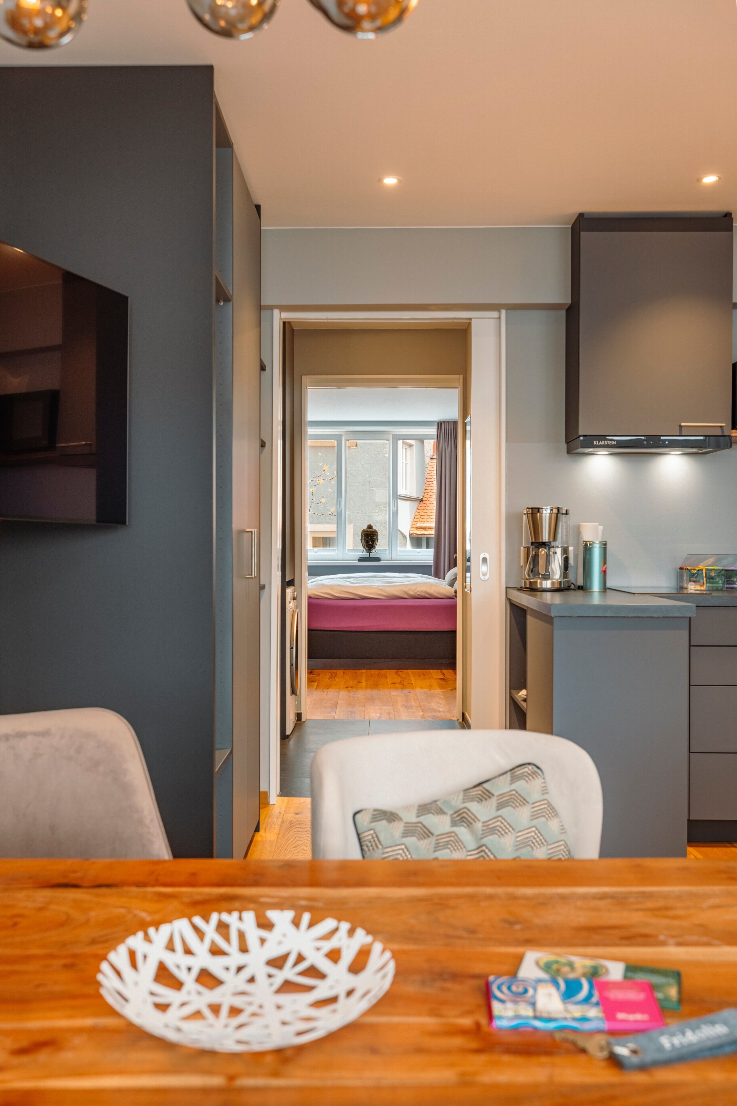 Una camera d'albergo moderna con un tavolo da pranzo in legno, una TV a parete e vista sulla camera da letto adiacente.
