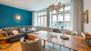 A modern living room with a sofa, dining table, and chairs, large windows, and a decorative mirror.