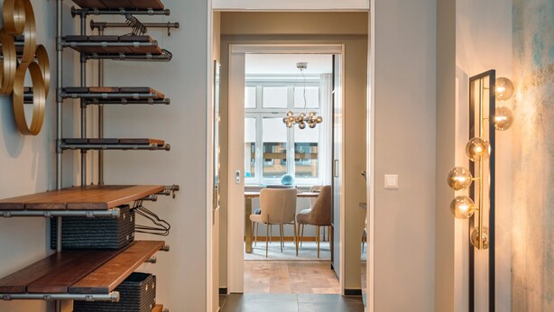 A modern hotel room with a bed, a wooden shelf unit, a mirror with lights, and a view into the dining area.