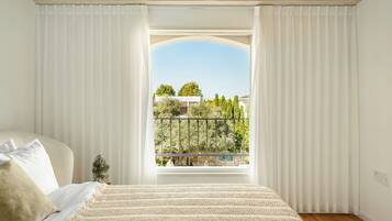 Un dormitorio con cama, cortinas blancas y vistas a un balcón con vegetación y barandilla.