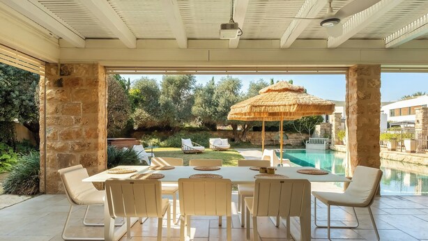 Un espace extérieur abrité, aménagé pour les repas, avec vue sur la piscine, des piliers en pierre et un parasol en chaume.