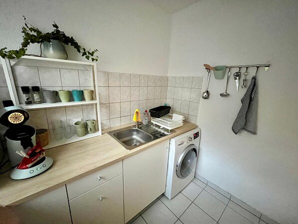 Una cocina con lavadora, un lavamanos y una estantería con diversos objetos.