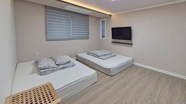 A small, modern room with two single beds, a TV mounted on the wall, and a window with blinds.