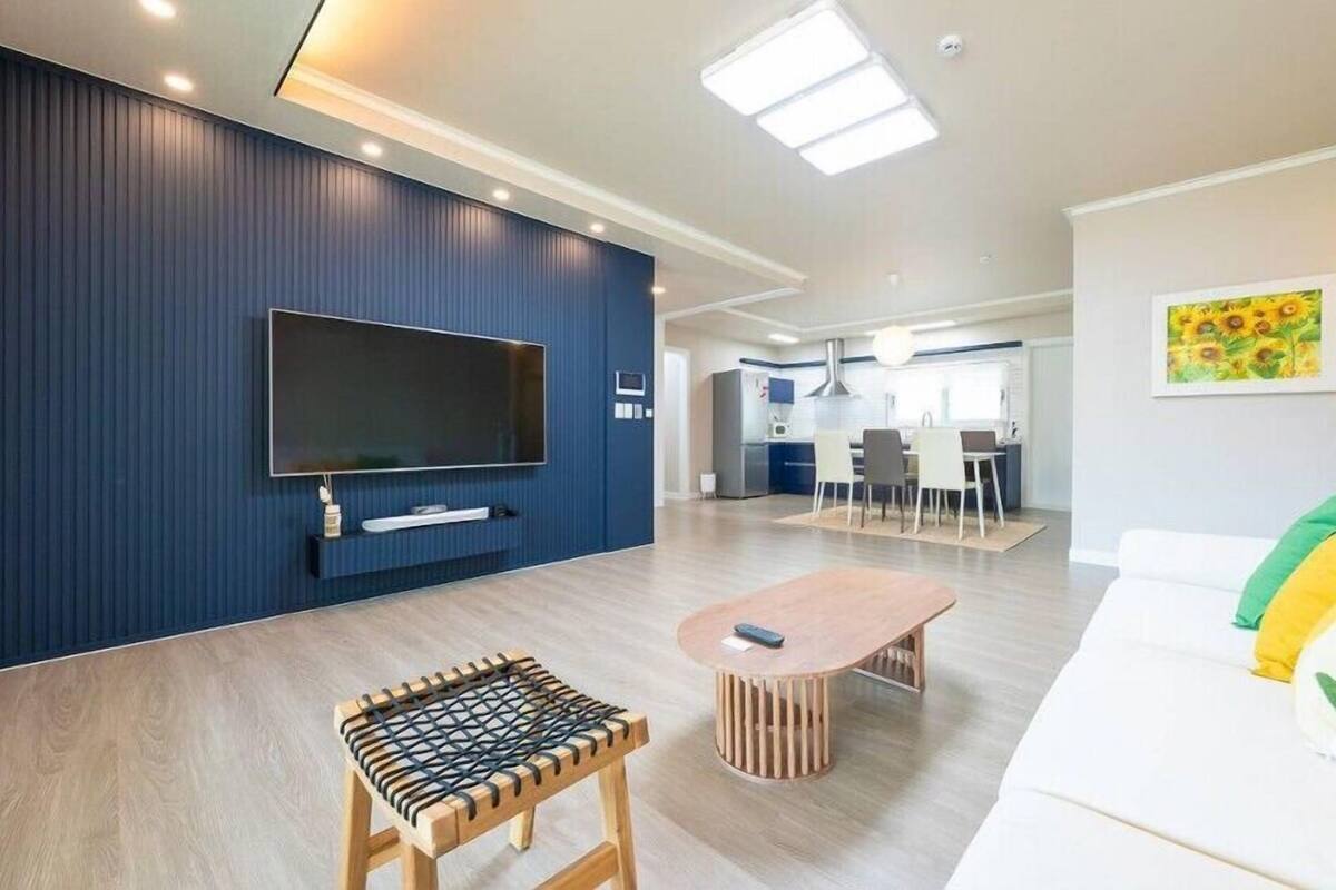 Ein modernes Wohnzimmer mit Flachbildfernseher, einer Holzbank, einem runden Couchtisch und einem weißen Sofa mit bunten Kissen.