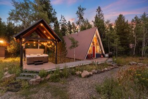 A cabin with a hot tub and a deck, surrounded by trees and a gravel path.