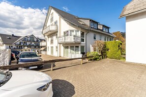 Ein Wohngebäude mit Balkon und einer gepflasterten Auffahrt, davor ein geparktes Auto.