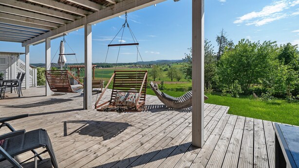 Un terrazzo in legno con mobili da esterno, una sedia a dondolo e vista su un paesaggio verde.