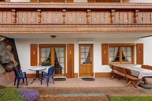 A building with a balcony, wooden doors, a round table with chairs, and a bench.