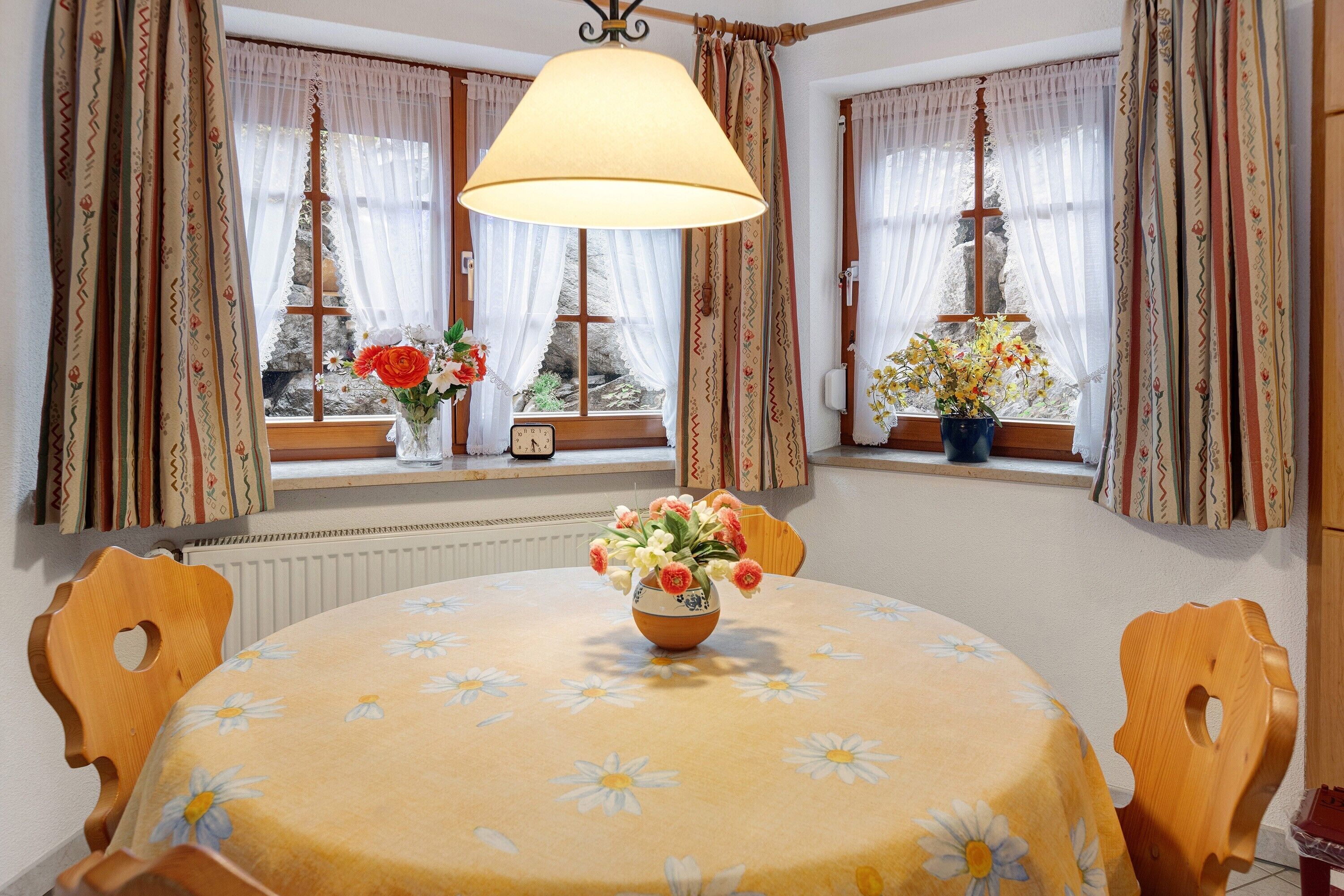 Un comedor con una mesa dispuesta para dos, un jarrón con flores y cortinas en las ventanas.