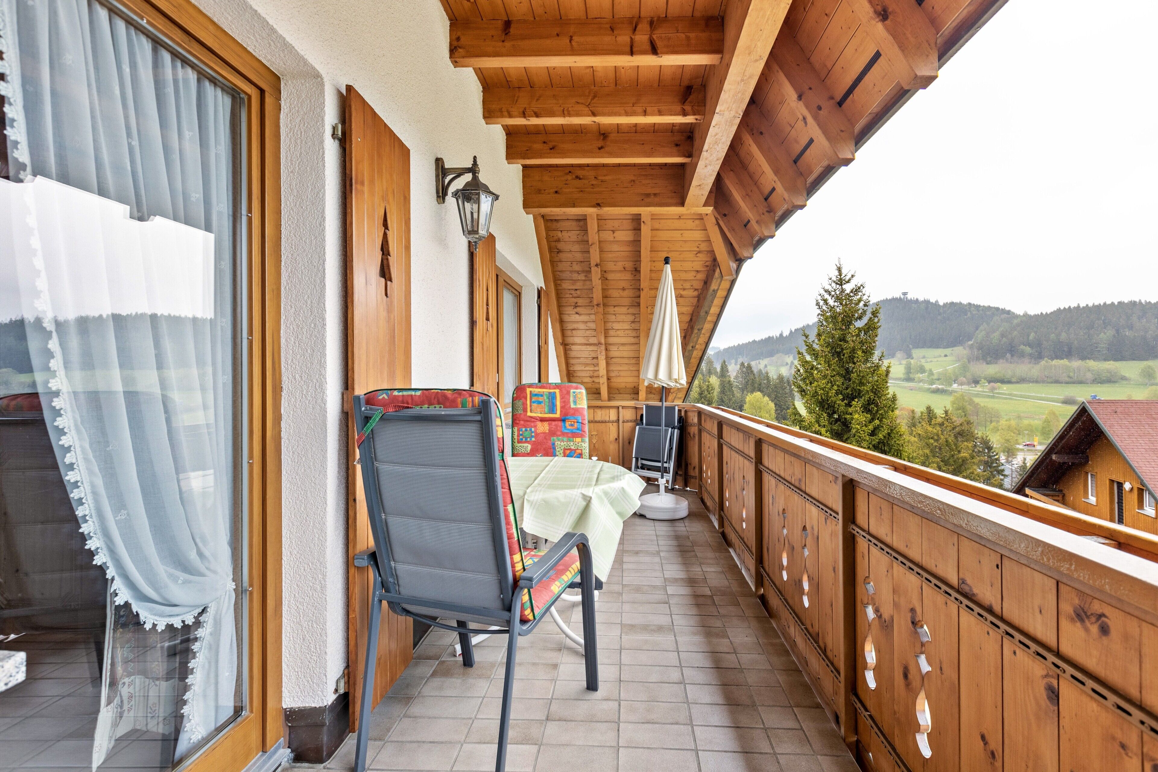 A wooden balcony with a table and chairs, a view of the countryside, and a wooden railing.