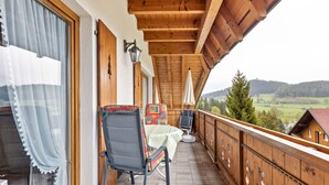 A wooden balcony with a table and chairs, a view of the countryside, and a wooden railing.