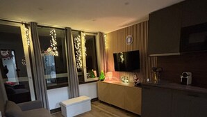 Uma sala de estar moderna com um sofá, uma pequena mesa branca, uma TV na parede e uma área de cozinha com bancada e armários.