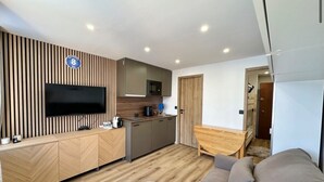 Uma sala de estar moderna com um sofá, uma televisão de tela plana presa a uma parede de madeira, uma cozinha com micro-ondas e piso de madeira.