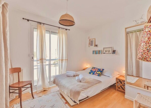 A bedroom with a bed, a chair, a bedside table, a mirror, and a window with curtains.