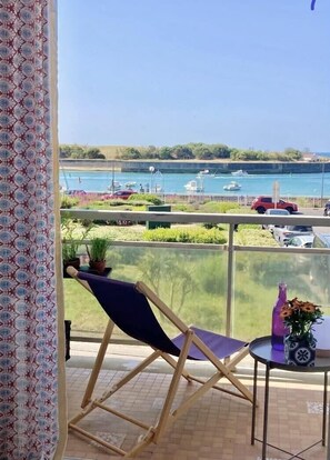 A balcony with a view of a marina, a chair, a small table with a vase, and a patterned curtain.