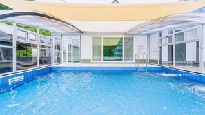 En swimmingpool med kristallklart blått vatten, omgiven av glasväggar och fönster, och ett solskyddande tak ovanför.