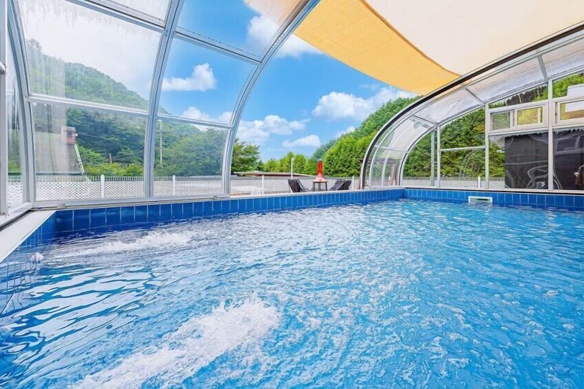 An indoor swimming pool with clear glass walls, a blue tiled floor, and a view of greenery outside.