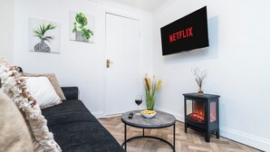 A modern living room with a black sofa, a round coffee table with a bowl and wine glass, a TV displaying 'NETFLIX', and a wood-burning stove.