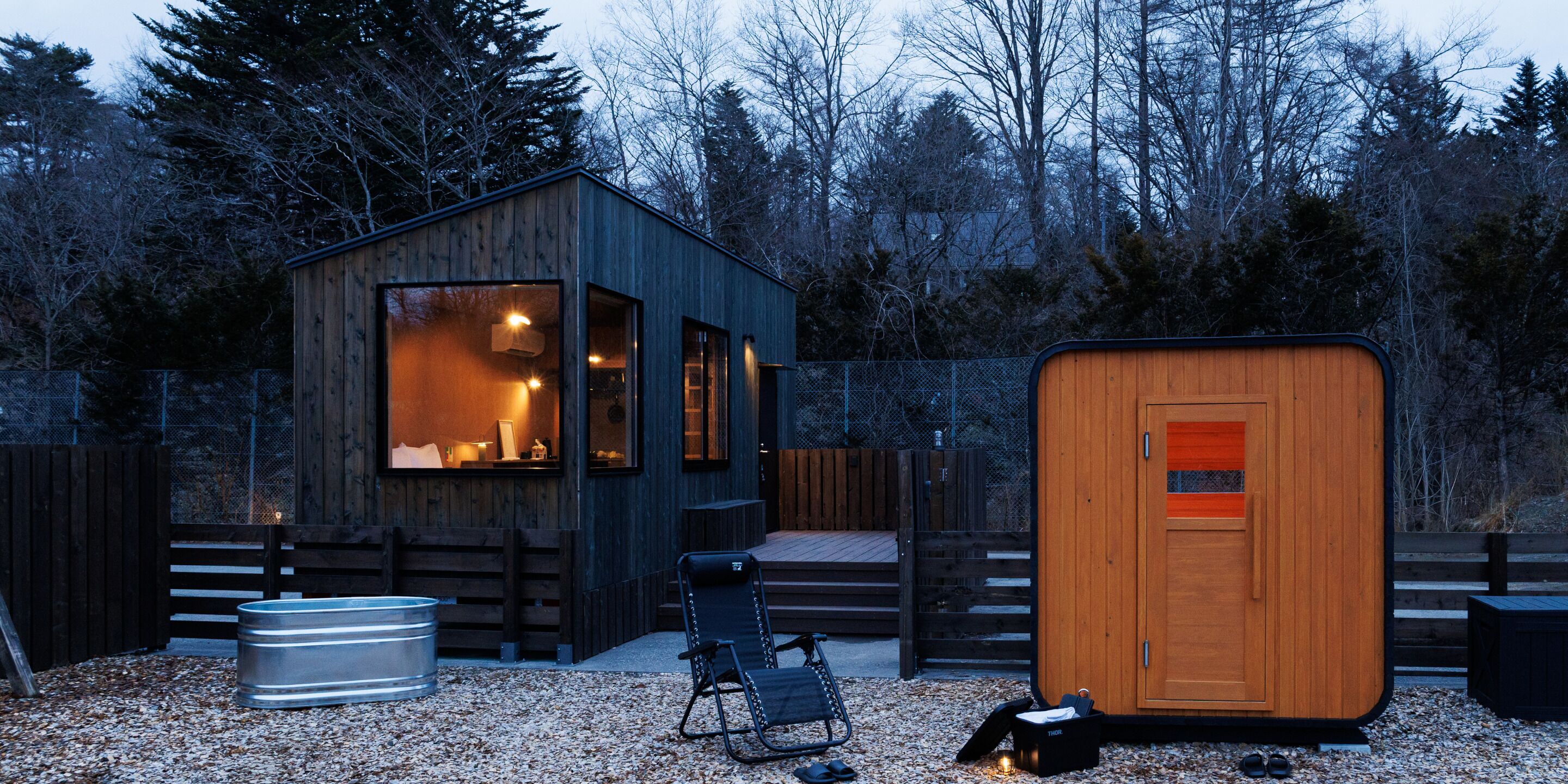 A cabin with a large window, a wooden door, and a metal barrel.