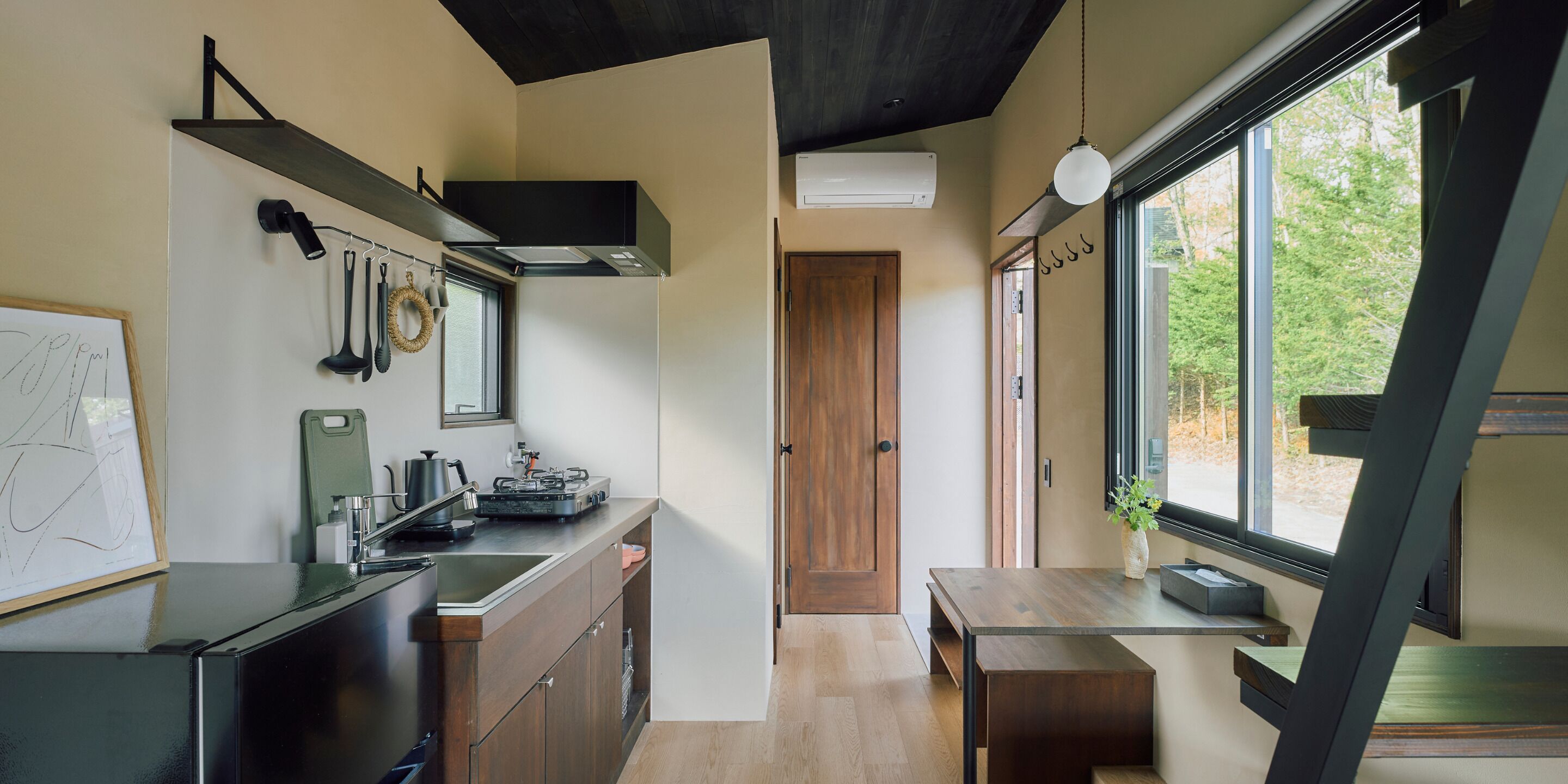 Eine moderne Küche mit einer Holzausziehtheke, einer Spüle und einem Herd. Eine Holztür führt in einen weiteren Raum, und ein Fenster bietet Blick auf Bäume.