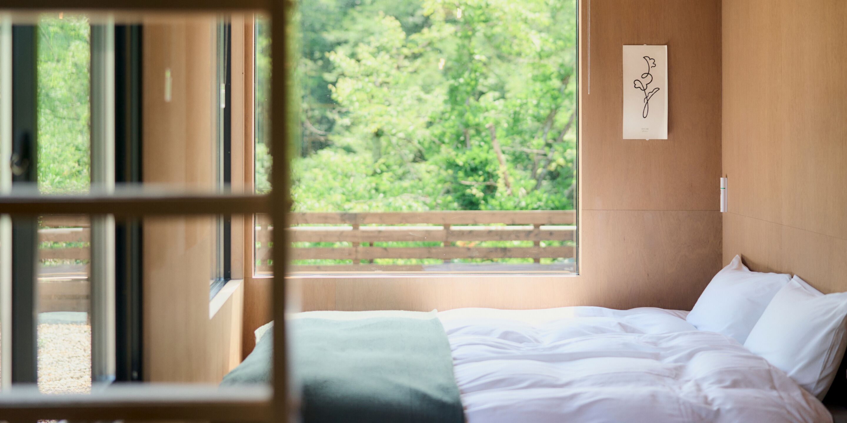 A room with a bed, a window with a view of greenery, and a wall-mounted artwork.