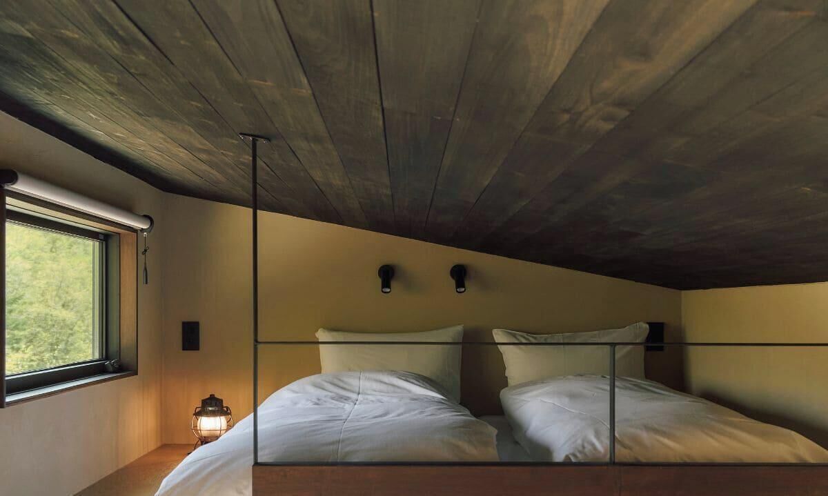 A room with a wooden ceiling, a bed with white bedding, a window with a view, and a wall-mounted light fixture.