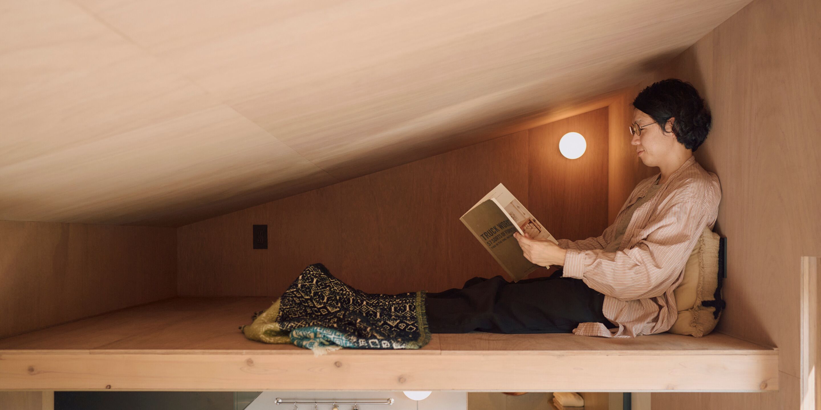 A person sitting on a wooden bunk bed reading a book in a room with wooden walls and a sloped ceiling.