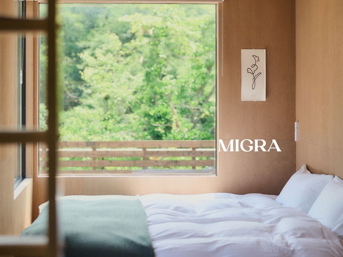 A bedroom with a large window, a bed, a wall-mounted artwork, and the word 'MIGRA' visible.