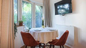 A dining area with a round table, two chairs, a TV mounted on the wall, and a window with curtains.