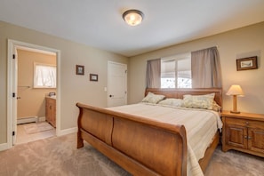 A bedroom with a wooden bed, a nightstand with a lamp, a window with curtains, and a door leading to another room.