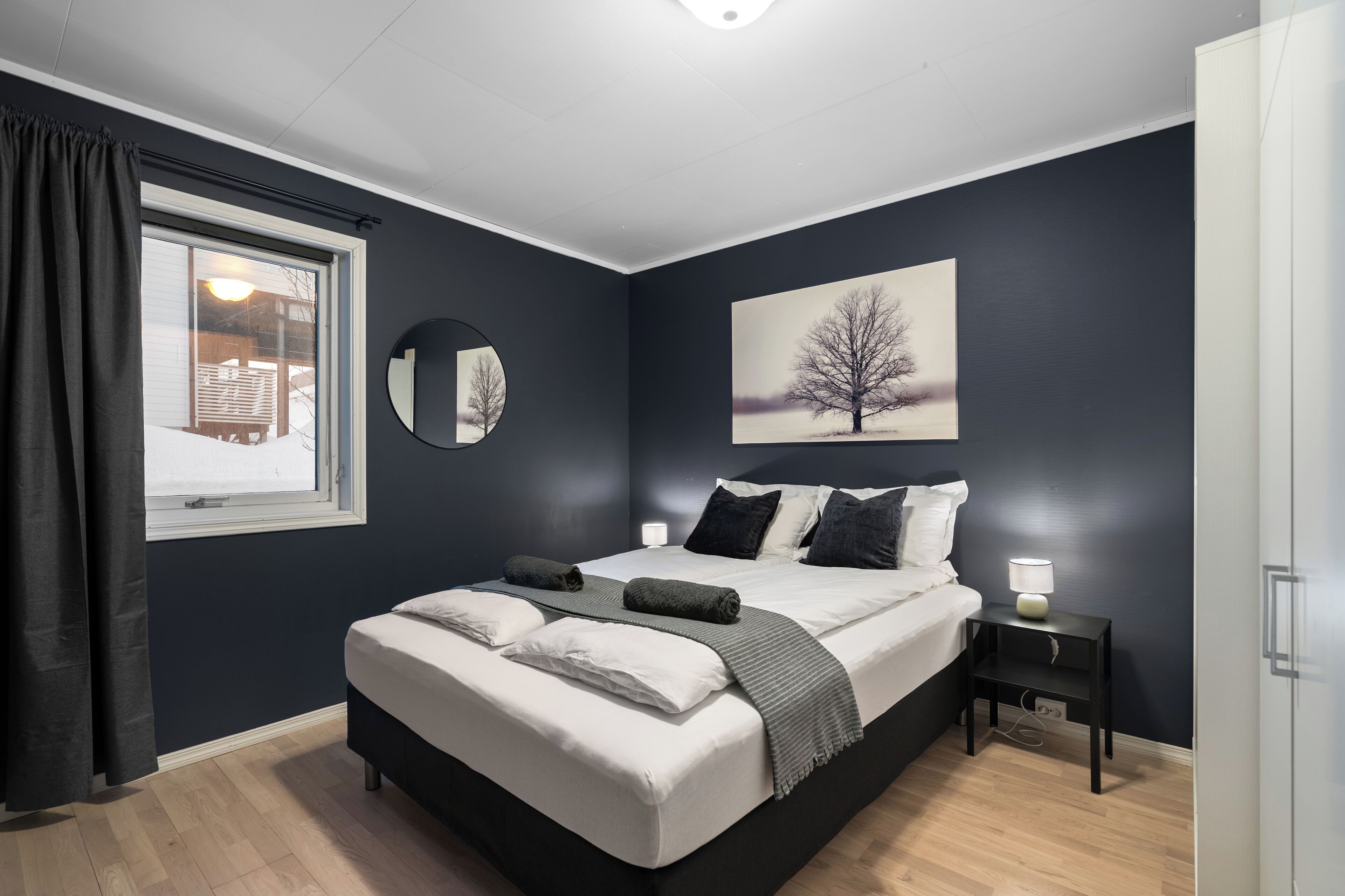 Une chambre à coucher avec un grand lit, une table de chevet avec une lampe, une fenêtre avec des rideaux et un miroir fixé au mur.