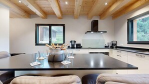 Une cuisine moderne dotée d’un îlot central, de poutres en bois au plafond et de grandes fenêtres offrant une vue sur l’extérieur.