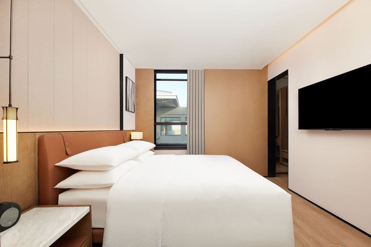 Ein modernes Hotelzimmer mit einem großen Bett, einem flachbildfernseher an der Wand, einem Nachttisch mit Lampe und einem Fenster mit Blick auf die Außenansicht des Gebäudes.
