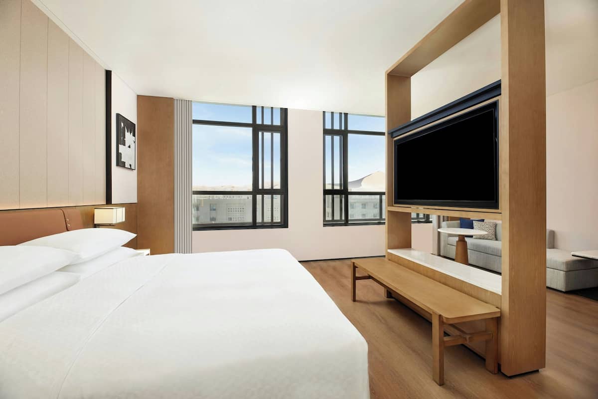 Ein Hotelzimmer mit einem großen Bett, einem Fernseher, einer Bank und großen Fenstern mit Blick auf die Stadt.