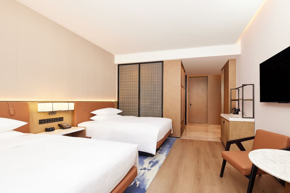 Ein Hotelzimmer mit zwei Betten, einem Schreibtisch, einem Stuhl, einem Fernseher und einer kleinen Küchenzeile.