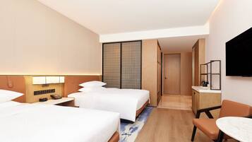 A hotel room with two beds, a desk, a chair, a TV, and a small kitchenette.
