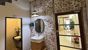 A bathroom with a sink, mirror, and toilet, a brick wall, and a glass door leading to a dining area.